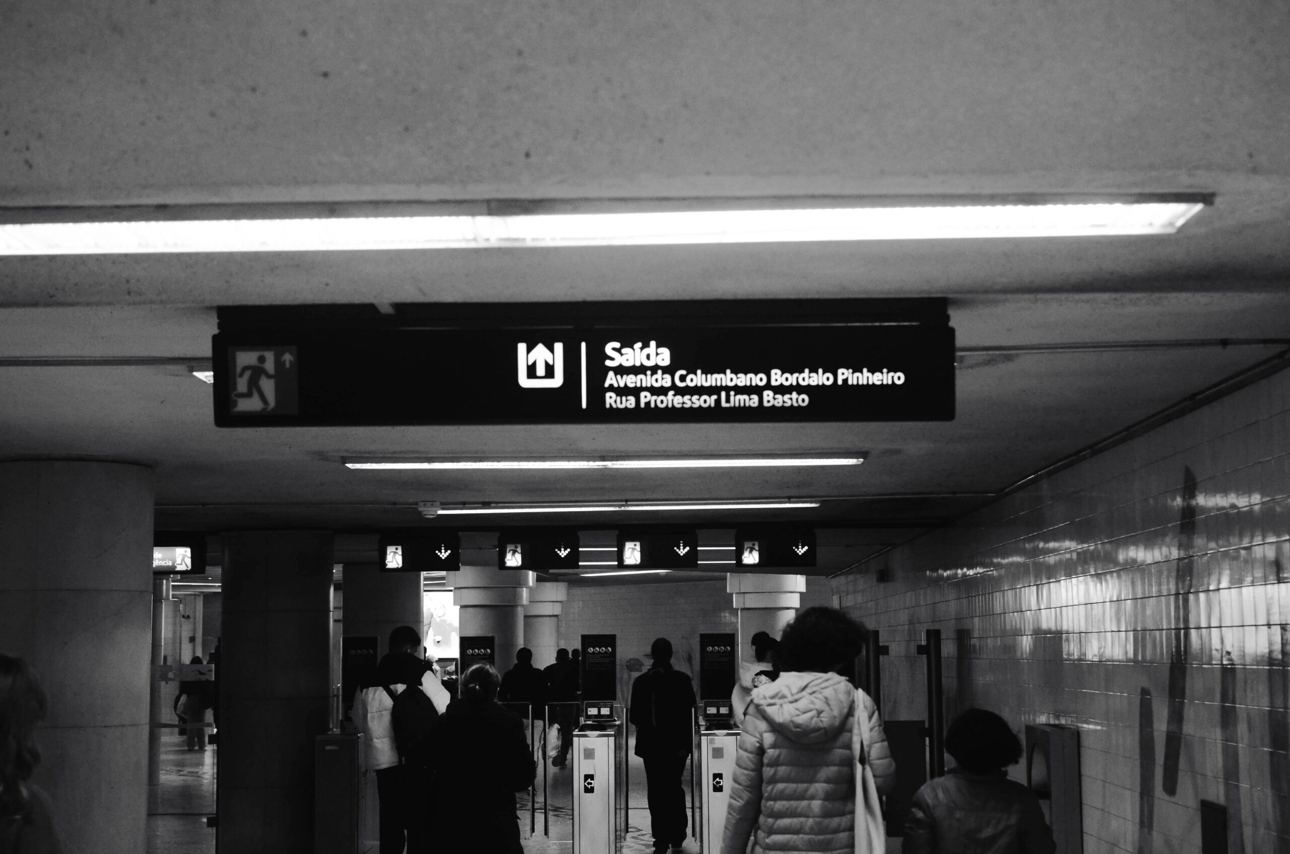 Monochrome photo of a subway station exit sign with people walking towards turnstiles.